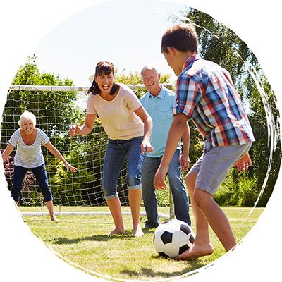 A multi-generational family playing soccer in a sunny backyard; a boy dribbles toward the goal as adults cheer.